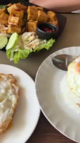 The video shows a dining table with a meal laid out. There is a plate of fried tofu with garnishes, a fried egg on a small plate, and a small container of sauce. Two people are visible. One is wearing a red top and black shorts, and the other is wearing black shorts. One of them picks up a fork from a napkin. The table is covered with a patterned placemat.