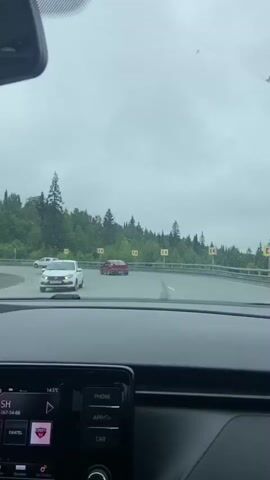 The video, shot from inside a car, shows a red car driving on a wet road through a wooded area. The red car is seen making a sharp turn. There are yellow directional signs on the side of the road. The car's dashboard display shows the time as 10:34 and the temperature as 14.5 degrees Celsius. The screen also displays music information, with the artist name "Yuldash" and a song title. Another white car is parked on the left side of the road. The sky is overcast.