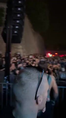 A night-time outdoor event is captured with a crowd of people visible. In the foreground, the back of a man's head and shoulders are visible, wearing sunglasses. Behind him, a large speaker stack is on the left. The crowd extends into the background, with a person in a white outfit standing on a raised platform, illuminated by a spotlight. Red lights are visible in the distance. Two men are seen interacting with a large piece of white fabric, possibly setting up or taking down a tent or screen. The environment appears to be a festival or concert setting.
