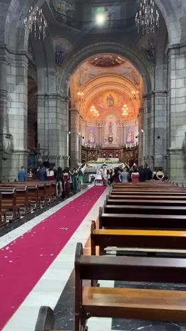 The video shows a wedding ceremony taking place inside a grand church. The perspective is from the back of the church, looking towards the altar. Pews are visible on both sides, with a red carpet runner leading up the aisle. At the front, a bride in a white dress and a groom in a light-colored suit stand facing each other at the altar, surrounded by a wedding party and guests. Many guests are holding up their phones to record or take pictures of the ceremony. The church interior features stone columns, arched ceilings with painted murals, and ornate chandeliers. The overall atmosphere appears to be a formal and joyous occasion.