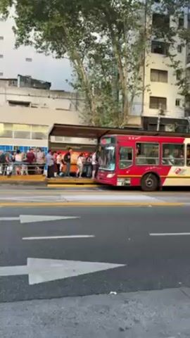 The video shows an outdoor urban scene. People are waiting to board a red bus at a station. There are several individuals standing in line, and the bus is parked at the curb, with its doors open, allowing passengers to board. The station has a covered shelter. The street has directional markings with white arrows, indicating the flow of traffic. Across the street is a building with windows and a white façade. Trees are seen in the background. A woman is walking in the foreground.
