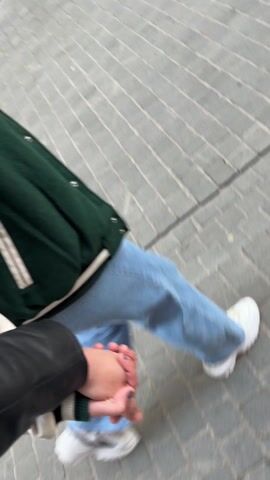 The video starts with a close-up of two people holding hands while walking on a paved surface. One person is wearing blue jeans, a white shirt, and a green varsity jacket with white accents. The other person is wearing a black leather jacket. The camera angle then shifts to a view of a street lined with multi-story buildings on both sides. Several people are walking along the street. The street is decorated with colorful, orb-like lanterns hanging overhead. The sky is a pale white or light gray, suggesting it's overcast or near dusk. There are shops and businesses along the street with signs and awnings.