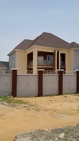 The camera pans across a row of single-story and two-story residential houses in what appears to be a developing or newly developed neighborhood. The houses feature fenced yards with decorative gates, mostly in shades of grey and white. The road is unpaved and dusty, appearing to be composed of sand and small rocks. The weather is clear and sunny with a bright, blue sky. The architectural style of the houses is modern with pitched roofs, and there is little visible landscaping. One house features a balcony. There are no people or vehicles visible. Some of the fences are topped with security features, possibly electric fencing.