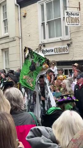 A street parade is in progress in front of a shop named Roberts Curios. A person in a large, white, bird-like mask, featuring a yellow beak and black eye patch, walks in the parade. The costume has black and grey fringe trailing from the body. A green flag emblazoned with an image and the words "RILEY'S CREW" is visible to the left of the costumed person. The street is crowded with spectators, some in costume.