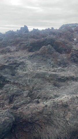 The video shows a rocky landscape near the ocean. The rocks are mostly gray and black, with some reddish hues. The sky is overcast. The ocean is visible in the background. There are no people or animals present in the scene. The video seems to be shot from a stationary position.