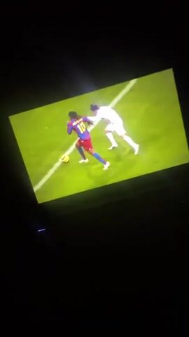 The video shows a laptop screen displaying a soccer match. The first shot is of a player in a Barcelona jersey, #10, dribbling past a Real Madrid player. The Barcelona player scores. The next shot is of a crowd in the stadium. The final shot is of the scoreboard showing Real Madrid 0, Barcelona 3. The lighting is low in the room, making it difficult to see the laptop's surroundings.