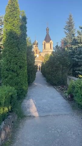 The video shows a stone pathway leading to a church with a dome and cross. There are trees and bushes on either side of the pathway. Two people are visible walking towards the church. The sky is clear and blue, indicating a sunny day. The video appears to be filmed from a low angle, looking up towards the church.