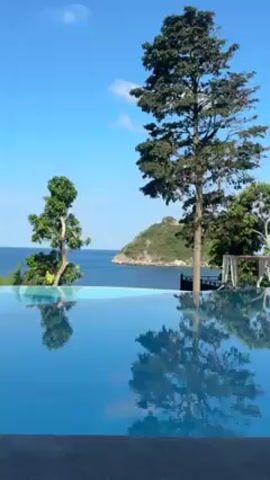 The video showcases a tranquil scene featuring an infinity pool overlooking a sea view. The pool's surface reflects the surrounding trees, cabanas, and a small island in the distance under a clear blue sky. There is no human presence or significant activity visible in the frame. The video captures the serenity of a tropical resort setting.