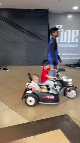In a well-lit, indoor space with a tiled floor, two young children are on a black, battery-powered motorcycle with a sidecar. The child driving the motorcycle is wearing a red shirt and light-colored pants. The younger child in the sidecar is wearing a striped shirt and light-colored pants. An adult male is walking alongside the motorcycle, holding onto the handlebars. A large black covering drapes down behind them, and a sign with the words "REDEFINE YOURSELF" is visible in the background. The motorcycle has silver accents and red details on the wheels. The adult is wearing a blue and black shirt and light blue jeans.