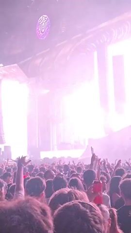 A concert crowd is visible from behind, with their hands raised in the air. On the stage, a DJ is performing, silhouetted against a backdrop of large screens displaying abstract visuals and lighting effects. Smoke or fog machines are creating a hazy atmosphere, and spotlights are illuminating the stage. The overall scene suggests a lively and energetic music event.