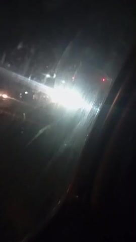 A car is seen driving on a road at night. The car is dark in color and appears to be a sedan. The headlights of the car are on and illuminating the road ahead. There are other vehicles in the background, also with their headlights on. The scene is dark, indicating that it is nighttime. The road appears to be paved.