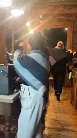 A woman in a blue and grey shawl and tan pants walks behind a counter to a black speaker. Another woman stands behind her, looking at a phone. A blonde woman walks toward an open door in the background. The first woman sets her hand on the speaker. The second woman then looks at the camera and smiles. The room has wooden walls, countertops, and cabinets. There is a Red Bull sign and a brass teapot on the counter. The video is a little shaky.