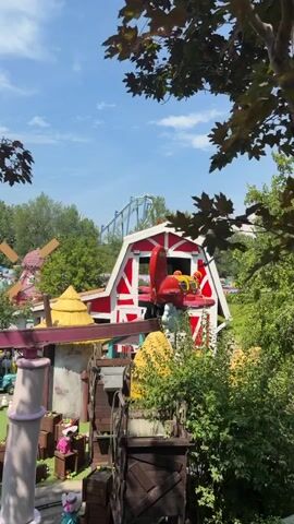 The video shows a children's amusement park ride with a red barn structure. A red airplane-shaped ride vehicle is on a track in front of the barn. Trees and foliage surround the ride. In the background, a roller coaster can be seen. The sky is blue with some clouds. The video appears to be filmed during the day.