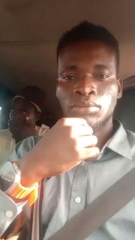 A video shows a person sitting in the driver's seat of a car. The person is wearing a gray button-down shirt, a seatbelt, and a smart watch with an orange wristband. They make the “peace” sign for the camera. Another person is sitting in the back seat of the car. The interior of the car is visible. The sun appears to be shining. Music is audible.