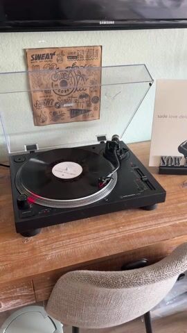 A slow panning shot reveals a turntable with a vinyl record playing on a wooden table. Next to it is a record album cover of Sade's "Love Deluxe," propped up on a small display stand. To the right is a large, black bookshelf filled with records, picture frames, and decorative objects. The room has pale green walls adorned with various posters and art prints. A white chair with a patterned pillow sits near a window in the background. The floor is light wood. The video provides a glimpse into a cozy, music-filled living space.