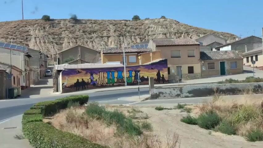 A time-lapse video shows a street scene in a rural town. A mural with colorful lettering and silhouettes of people and animals is painted on the side of a building. The sun is shining, and the sky is blue with a few clouds. The camera pans across the scene, showing houses, a road, and vegetation. The time-lapse nature of the video captures the subtle shifts in light and shadow as the sun moves across the sky.