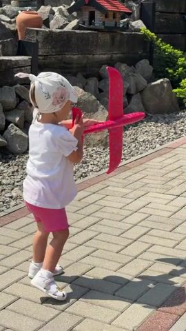 A young child, seen from behind, is playing outdoors in Wisła, Poland. The child wears a white short-sleeved top with puffed sleeves, pink shorts, white sandals, and a light-colored bandana or head covering on their head. Their light-colored hair is tied up.

The child is actively engaged in throwing a bright pink toy airplane. At the start of the video, they throw the airplane into the air. It briefly flies before landing on the paved ground. The child then moves to retrieve the airplane, picks it up, and throws it again.

The setting is an outdoor area, likely a garden or backyard. The ground is covered with a neatly paved walkway made of grey and red interlocking bricks in a herringbone pattern. To the left of the walkway, there is a landscaped area featuring numerous rocks and stones of varying sizes, along with green plants and shrubs. A small, decorative wooden structure with a red roof is visible among the rocks. The scene is brightly lit by sunshine, casting distinct shadows of the child and the airplane on the ground. No text is visible in the video.