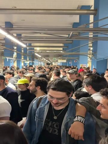 The video shows a dense crowd of people in what appears to be a subway station or underground walkway. The space is brightly lit with fluorescent lights attached to a grid of overhead pipes. The walls are painted a light blue color. The people are diverse in age and appearance and are dressed in casual clothing. Many individuals are wearing hats or caps. There is little visible movement, suggesting the crowd is relatively stationary and compacted. One man in the foreground is wearing a denim jacket over a black t-shirt and has a necklace. He is smiling slightly and has glasses on. Another man beside him has his arm around his shoulder. A bracelet is visible on the man's wrist that reads "KANYE". The environment appears to be indoor.