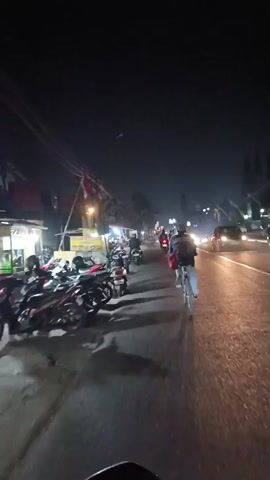 The video shows a nighttime street scene. The perspective appears to be from a moving vehicle, possibly a motorcycle, looking forward. On the left side of the road, there are several parked motorcycles and a row of small food stalls or shops with illuminated signs. People are visible sitting at tables outside one of the establishments. To the right of the road, traffic is moving, with car headlights and taillights visible. A cyclist is also visible ahead, riding in the same direction as the viewer. The road surface appears wet, suggesting recent rain. The sky is dark and clear, indicating a nighttime setting.