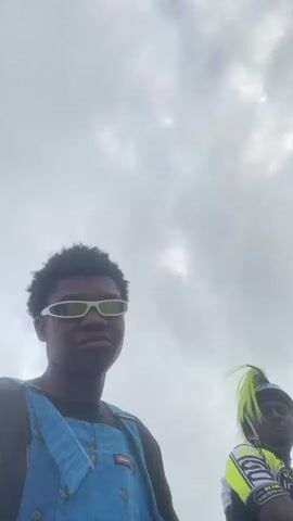 The video begins with a close-up of a young Black person wearing blue overalls over a black t-shirt and white futuristic sunglasses. They look directly at the camera with a neutral expression. In the background, slightly out of focus, another person is visible with bright neon green hair, wearing a black and yellow cycling jersey and red pants. They are sitting down on a rocky surface. As the video progresses, the camera pans slightly to the right, revealing more of the environment, which appears to be a rocky coastline with the ocean in the background. The sky is overcast. The person with green hair makes a peace sign with their fingers and smiles. A third person, a Black male with no shirt on, is visible sitting further to the right in the background. A green helmet is on the ground near him. The person in the overalls raises their hand, showing their fingers, then lowers it. The person with green hair continues to make gestures and smiles. The video ends with a shot of the ocean and the rocky shore.