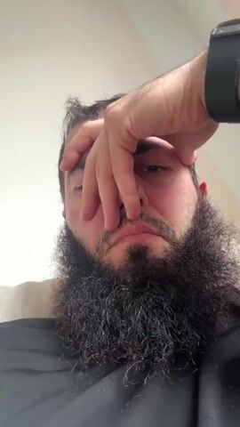 A close-up, low-angle shot shows a man's face from the nose down, with his hand covering his eyes. The man has a long, thick beard. He is wearing a black garment and a black smartwatch on his left wrist. The background is blurred and appears to be a light-colored wall and furniture.