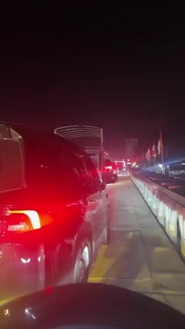 The video depicts a line of vehicles at night, likely on a bridge. The camera is positioned inside a car, capturing the rear view of the car in front. The car's taillights are brightly illuminated, casting a red glow on the surrounding area. Several Indonesian flags are visible along the side of the bridge, illuminated by lights. The overall environment appears dark, and the scene suggests a standstill or slow-moving traffic situation at night.