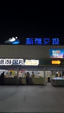 A nighttime exterior shot shows a service area with several stalls and shops. In the foreground, two people are walking toward the camera. The person on the left wears a red t-shirt and white shorts, and waves. The person on the right wears a white t-shirt and white shorts, and holds a purple object to their mouth. Behind them, a food stall called "Road Tour Bazaar" is visible, with several people working behind the counter. Above the stalls, a large illuminated sign reads "新 疆 投" in blue letters. To the right of this sign is a digital billboard featuring a man and the text "看过欧莱雅 再来找我" in Chinese. Further to the right, another illuminated sign reads "石河子服务区" in red letters. Various other signs for businesses like "JVC" and "Cotti Coffee" are also visible. The ground is paved, and the overall lighting suggests it is evening or night.
