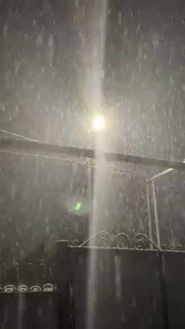 The video shows a heavy snowfall at night, illuminated by a bright street lamp. A dark fence with decorative swirls along the top edge is visible in the foreground. Above the fence, a simple metal structure, possibly a clothesline or support frame, is also visible against the backdrop of falling snow. The street lamp casts a strong, vertical beam of light through the snow, highlighting its intensity. There are no people or interactions visible in the video. The environment is characterized by the darkness of the night and the heavy snowfall, creating a stark and cold atmosphere.