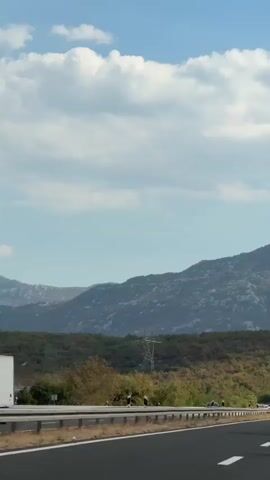 This video shows a view from a car traveling on a highway. The highway is paved with asphalt and has white lane markings. On the left side of the highway, there is a guardrail and then a landscape with trees and vegetation. In the background, there are mountains. The sky is blue with some white clouds. There are other cars on the highway, some of which are visible in the distance. The sun appears to be shining.