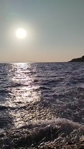 The video shows a close-up view of the ocean on a sunny day. The sun is high in the sky, casting a bright reflection on the water. Small waves are gently lapping at the shore, creating a peaceful and serene atmosphere. The water is a deep blue, and the sunlight creates a shimmering effect on its surface. The scene is calm and tranquil, evoking a sense of relaxation and natural beauty.