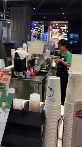 The video shows a young woman in an apron and hairnet working behind a counter at what appears to be a beverage stand or cafe. She is standing to the right of the frame, facing left, and is holding a small metal pot or cup in her hands, appearing to be preparing a drink. To her left, there is a stainless steel counter with various items, including a black coffee machine with a white cloth draped over it, a red bottle, and a clear dispenser. In the foreground, stacks of white paper cups are visible, some with green animal logos and others with patterned designs. There is also a menu or promotional display on the left side of the frame with images of drinks. The background shows a dimly lit retail environment with displays and signage, suggesting a shopping mall or similar commercial space. The overall impression is of a busy service environment.