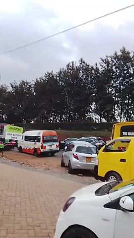 The video shows a traffic jam on a road. There are several cars and buses, including one with the words "THE MARION SCHOOLS" written on it. The buses are yellow.  Trees are visible in the background. A person walks in front of the camera holding what appears to be a phone. There is also a billboard above the road. The sky is overcast.