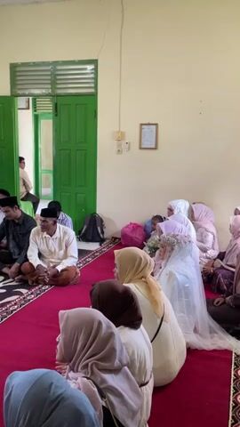 The video shows a gathering of people, mostly men, sitting on the floor on a red carpet with a patterned rug in the center. The room appears to be indoors, with light green walls and a white ceiling. There are two green doors visible in the background, one slightly ajar, revealing another room with a man standing. A window with green curtains is also visible. Some individuals are wearing traditional Islamic attire, including caps and robes. Towards the right side of the frame, a bride wearing a white wedding veil and dress is seated, accompanied by other women also in modest attire. The overall atmosphere suggests a formal event, likely a wedding ceremony or reception, given the presence of a bride and the gathering of guests. Some are engaged in conversation, while others appear to be listening or observing. Objects such as bags, books on shelves, and a small table are also present in the environment.