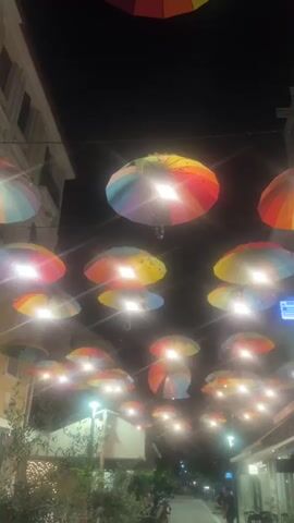 The video captures an outdoor street scene at night in Karditsa, Greece. Numerous colorful umbrellas, predominantly rainbow-colored, are suspended above the street, acting as decorative lighting. Many of these umbrellas have lights positioned beneath them, creating a bright glow and illuminating the area. The street below is lined with multi-story buildings, some of which appear to be shops or establishments. Streetlights are also visible on the sides. In the distance, a few people can be seen walking on the street. The overall ambiance created by the illuminated umbrellas is lively and festive. Text on signs on the buildings in the background is visible but not legible due to distance and lighting conditions.