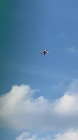 The video shows a red and white airplane performing aerobatic maneuvers against a blue sky with scattered white clouds. The airplane is small and appears to be remotely controlled or a small single-engine aircraft. The camera angle is looking upwards at the airplane, which is the sole subject of the video. The airplane is seen in various orientations as it performs loops and turns, sometimes appearing upside down and other times banking sharply. The video is short, lasting only a few seconds, and captures the dynamic movement of the aircraft in flight.