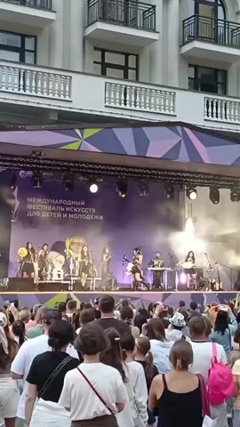 A live musical performance is taking place on an outdoor stage. A band, comprised of several female musicians, is actively playing instruments and singing. The stage is decorated with a large banner that reads "МЕЖДУНАРОДНЫЙ ФЕСТИВАЛЬ ИСКУССТВ ДЛЯ ДЕТЕЙ И МОЛОДЕЖИ" and "РОЗА ХУТОР." Lighting equipment is visible above the stage, and smoke effects are being used. In front of the stage, a crowd of spectators, including adults and children, are gathered, many of whom are holding up their phones to record the event. The background features a multi-story building with balconies.