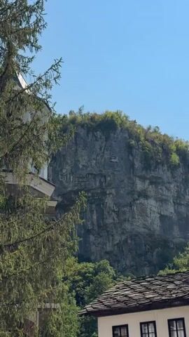 A vertical video shows a rocky cliff face with some trees in the foreground. On the cliff face, there is a carved symbol that appears to be in Greek letters. There are also some buildings visible in the lower portion of the video, suggesting a settlement near the cliff. The sky is clear and blue.