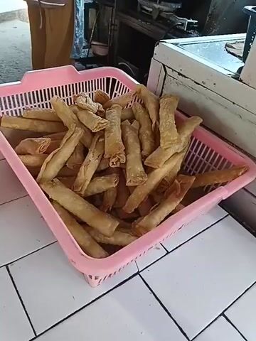 The video shows a close-up view of a pink plastic basket filled with fried spring rolls. The basket is placed on a tiled surface. To the left of the basket is a brown cardboard box, and next to the box is a clear plastic bag containing more spring rolls. The background is blurred but appears to be an outdoor or semi-outdoor setting with some shelving and other items. The lighting is natural and appears to be daytime.