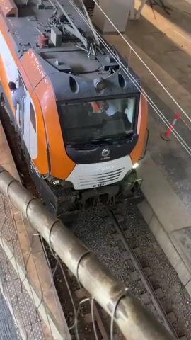 A long, orange and white passenger train sits at a station platform. The train has the branding "Alstom" and "ONCF". The train's number is E-1451. One man in a light-colored shirt stands in an open doorway of the train's front engine. Inside the front engine, a second man wearing an orange safety vest is visible. Passengers are standing and walking along the platform.