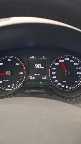 The video shows a first-person perspective from inside a car driving on a highway. The car's dashboard is visible, displaying a speed of 99 km/h. It is daytime, and the weather is overcast with light rain. The road is wet, and raindrops are visible on the windshield. Highway signs are visible overhead. The highway has multiple lanes and there is some traffic present. Trees and power lines border the highway on either side. The focus is on the road ahead, with the dashboard providing speed and vehicle information.