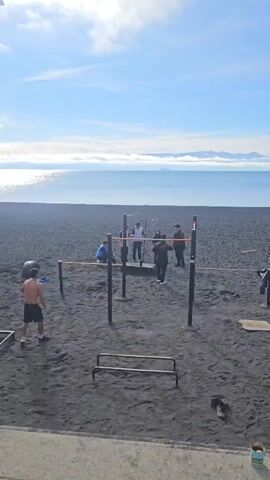 The video shows a group of people on a black sand beach next to a body of water under a partly cloudy, blue sky. In the background, distant mountains are visible. The scene includes several pieces of fitness equipment set up on the sand, including a pull-up bar and parallel bars. A shirtless man is walking toward the pull-up bar, while a group of people stand around the same equipment. Another person is walking towards the parallel bars. Two people are sitting at the bottom corner of the frame. A metal can sits on the ground beside them.