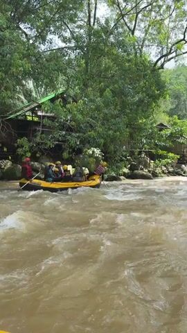 A yellow raft with several people wearing life vests and helmets navigates a churning, muddy river surrounded by lush green trees and vegetation. The people in the raft appear to be paddling. In the background, structures resembling huts or shelters are visible among the trees, with a bridge-like structure also present. The scene suggests an outdoor adventure activity in a tropical environment.