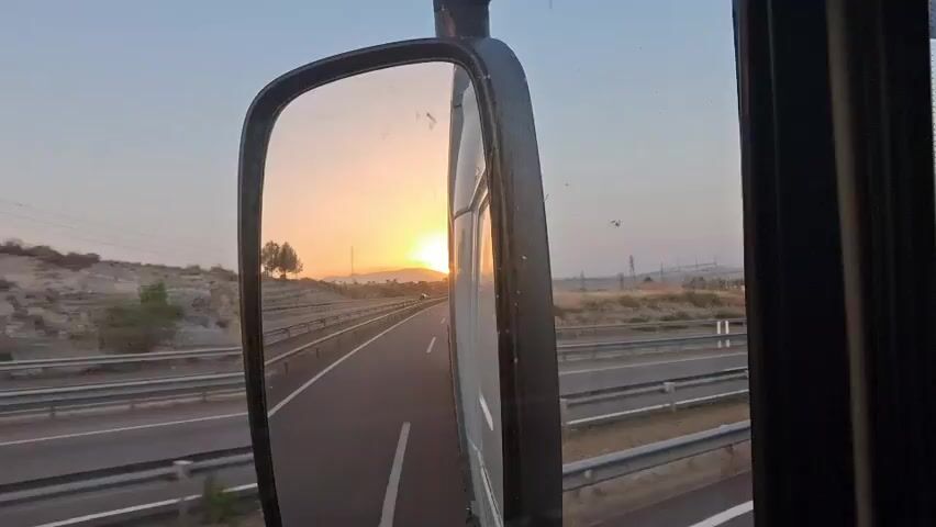 The video shows a view from inside a vehicle, looking into a side mirror. The mirror reflects a highway with a sunset in the background. The sun is low on the horizon, casting a warm glow over the landscape. The highway appears to be in a rural or semi-rural area, with hills and trees visible on either side. The vehicle is moving, as indicated by the motion blur in the reflection.
