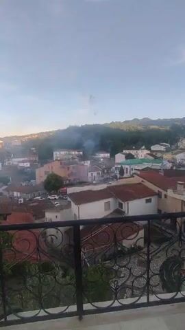 The video shows a view of a town with buildings and trees. There are flashes of light and smoke in the sky, indicating fireworks or explosions. The sky is clear with some clouds. The foreground shows a balcony railing with an ornate design.