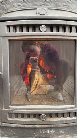 A young woman in a maroon sweater and dark tights is squatting within a cast iron wood-burning stove. She is holding a smartphone and taking a selfie. There is a split log positioned in front of her, and flames are visible around her. The stove has a glass door.