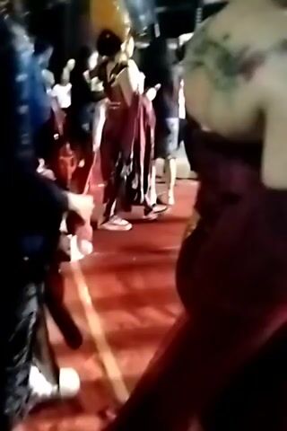 This is a dimly lit video that appears to capture a social gathering or event. In the foreground, a person wearing white sneakers and dark pants is visible, holding a red object. To their right, another person in a red garment is seen from the back, with a tattoo on their shoulder. The background is populated by other individuals, some of whom are in traditional attire, and illuminated by various lights, suggesting an outdoor or semi-outdoor setting with some decorations or structures. The overall impression is one of a lively event with people interacting and moving around.