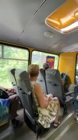 The video shows the interior of a yellow bus with several passengers. In the foreground, a woman with short blond hair wearing a floral dress is seated facing away from the camera. To her left, another person's legs are visible, indicating they are also seated. A poster with the word "ЦИРК" (Circus) in red letters is visible through a window. The bus has grey seats and a yellow ceiling with a skylight. In the middle ground, a person wearing a plaid shirt and a backpack is standing and appears to be interacting with someone through a window. Another person, an elderly woman, is seated to the right. The environment outside the bus appears to be green and treed.