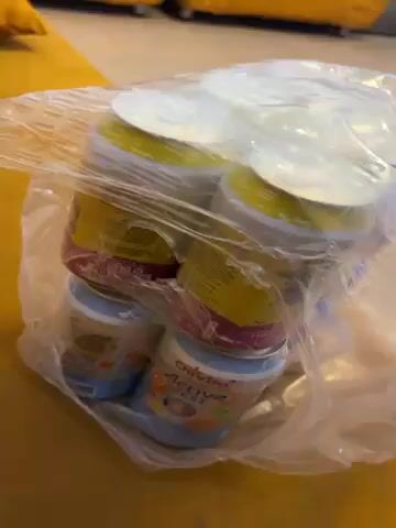 Here's a description of the video:

The video shows a clear plastic bag containing multiple containers of baby food. The containers are stacked on top of each other, with the top containers being yellow and magenta, and the bottom containers being blue. The containers appear to be unopened, with sealed lids. The bag is sitting on a yellow surface, possibly a couch or chair. There are yellow pillows visible in the background, slightly out of focus. The camera moves slightly, causing the bag and containers to shift in focus.
