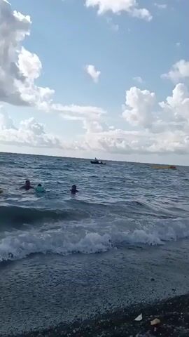 A view from the shore of a body of water with moderate waves and a cloudy sky. In the middle ground, a person is swimming, their head and shoulders visible above the water. Further out, a small boat with at least two people aboard is moving across the water, creating a wake. In the background, a yellow inflatable object is floating on the water. The scene appears to be a recreational area.