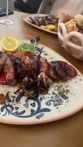 The video shows a man preparing to eat a meal. The table is set with a plate of grilled squid, lemon, green sauce, some roasted vegetables, and bread. Another plate with a similar dish is visible in the background. The man, wearing glasses and a beige sweater, squeezes lemon onto his food. A glass of white wine is also on the table. The scene appears to be in a restaurant setting.